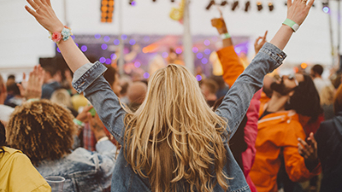 Young woman with blonde hair is dancing with her friends in a performance tent at a music festival. She has her arms outstretched and is watching the performance.
